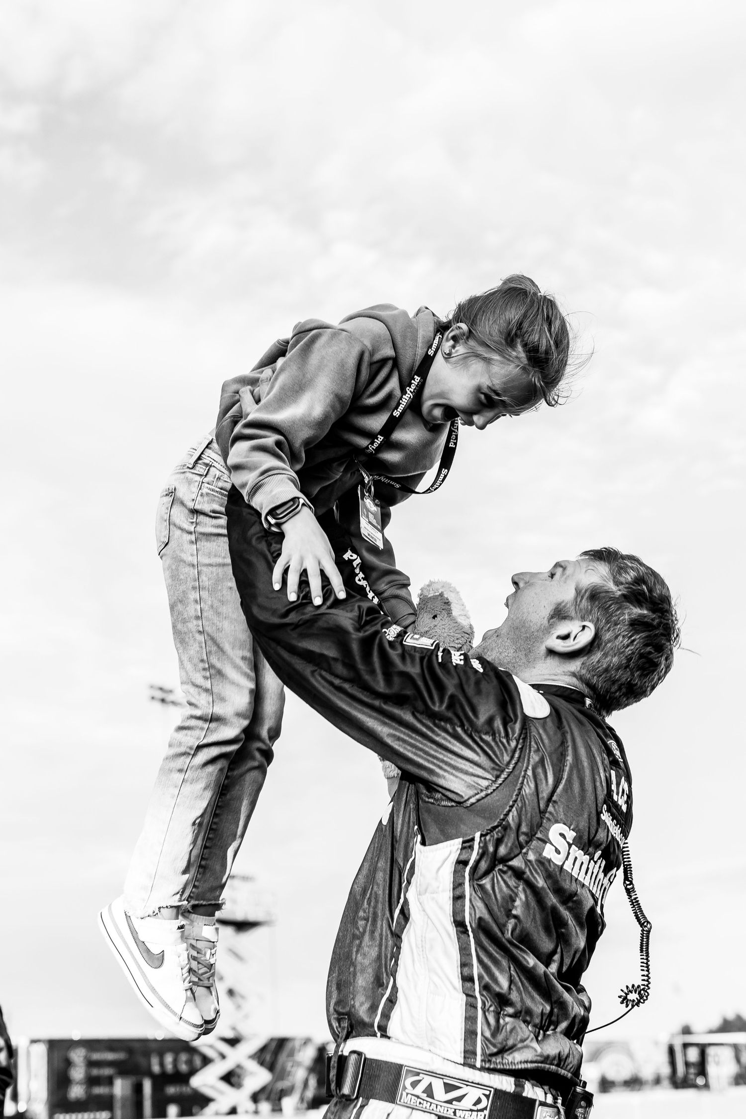 Man in racing suit lifting a child high in the air outdoors in black and white.