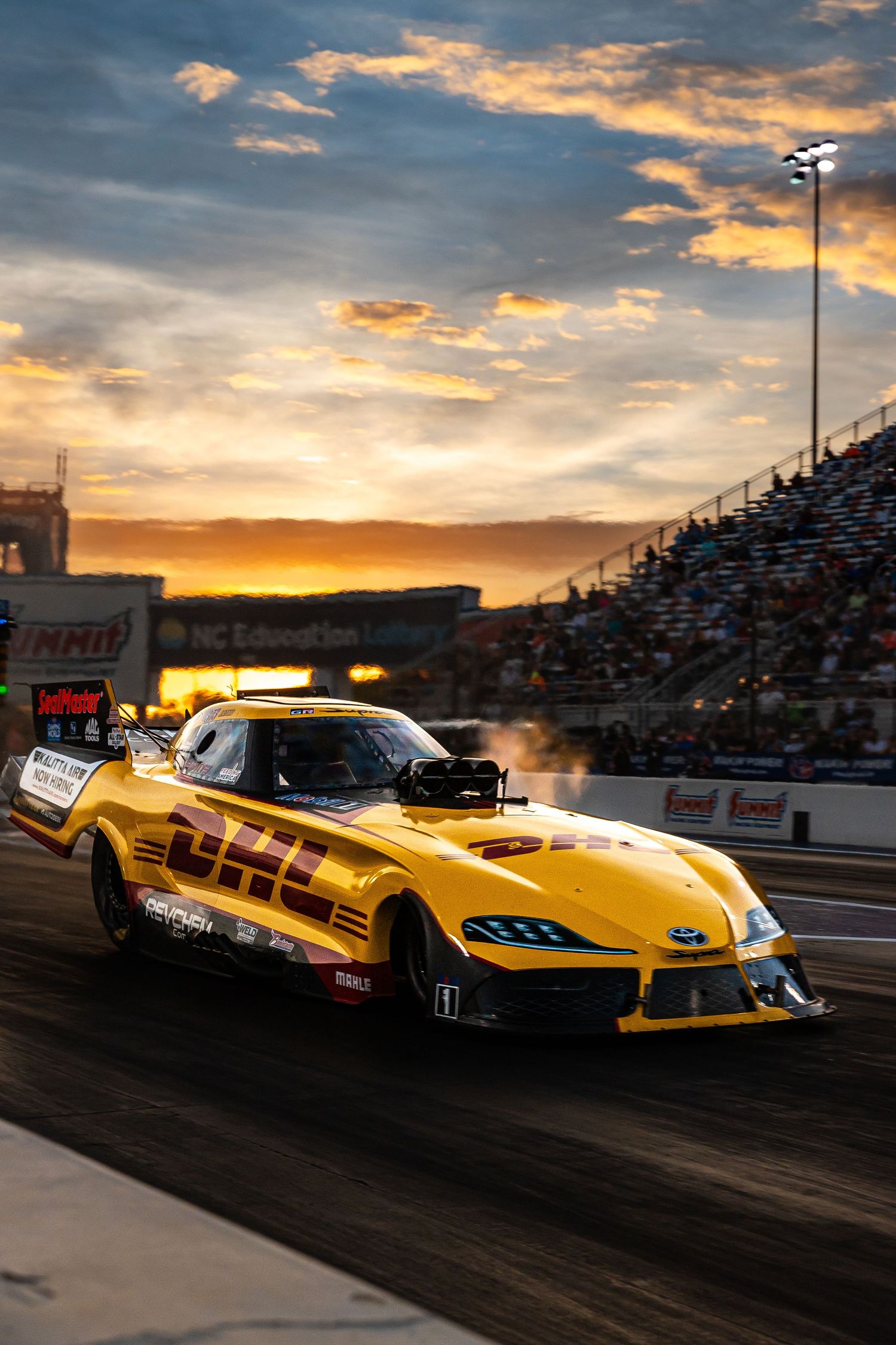 DHL-sponsored yellow funny car drag racing at sunset with dramatic orange sky and packed grandstands at NHRA event.