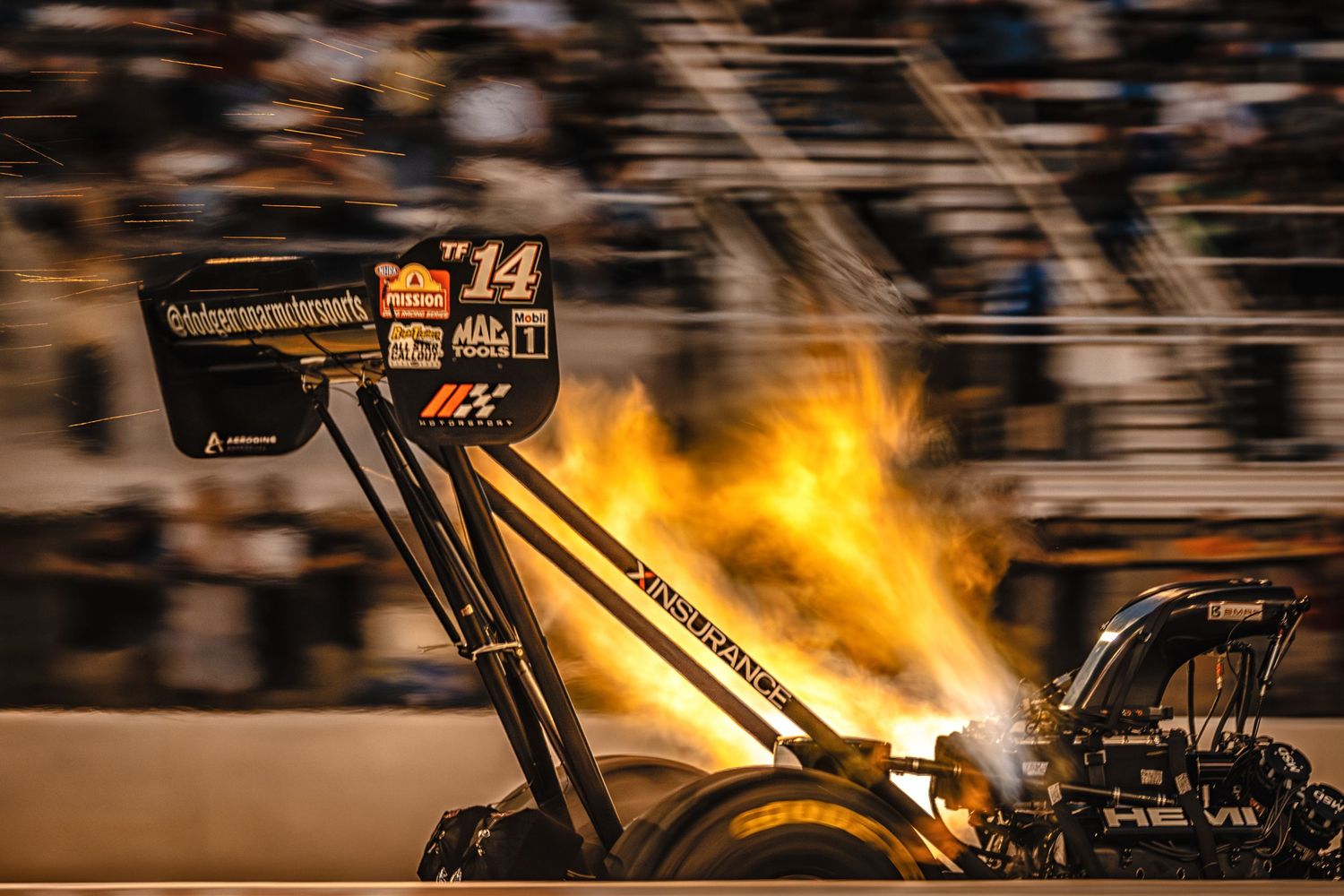 Top fuel dragster #14 with flames shooting from engine on racetrack at night during NHRA drag racing event.