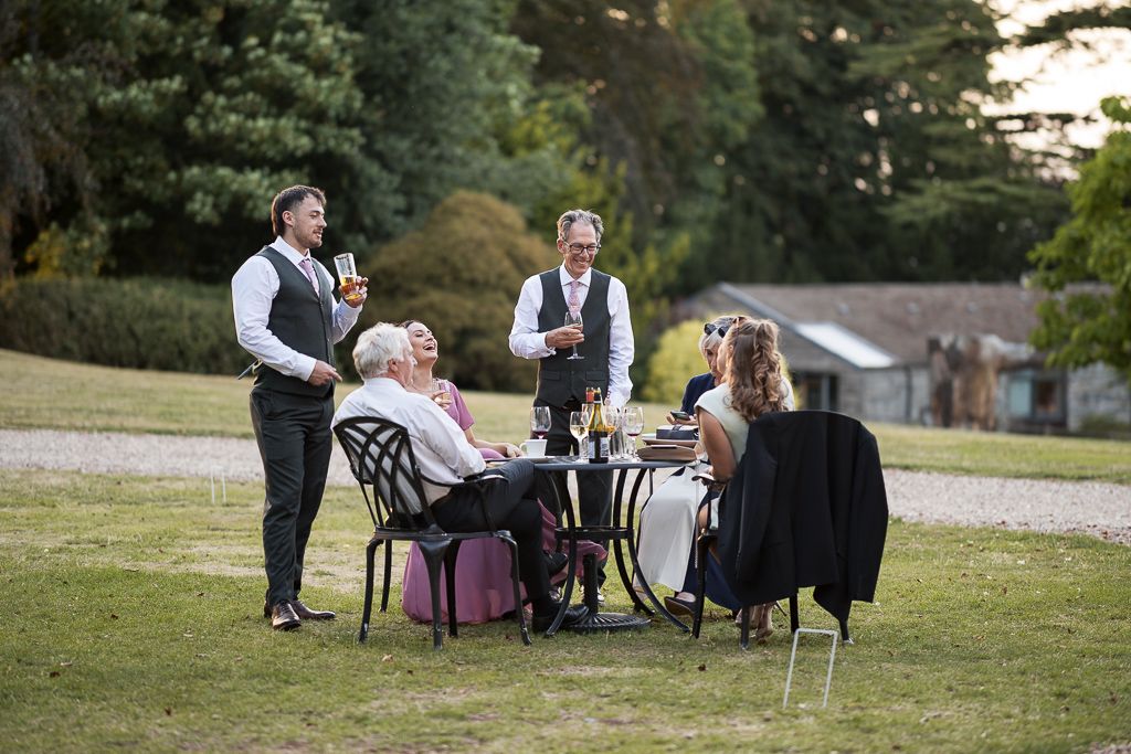 Elegant outdoor garden party with guests seated at a table being served by waitstaff in formal attire.