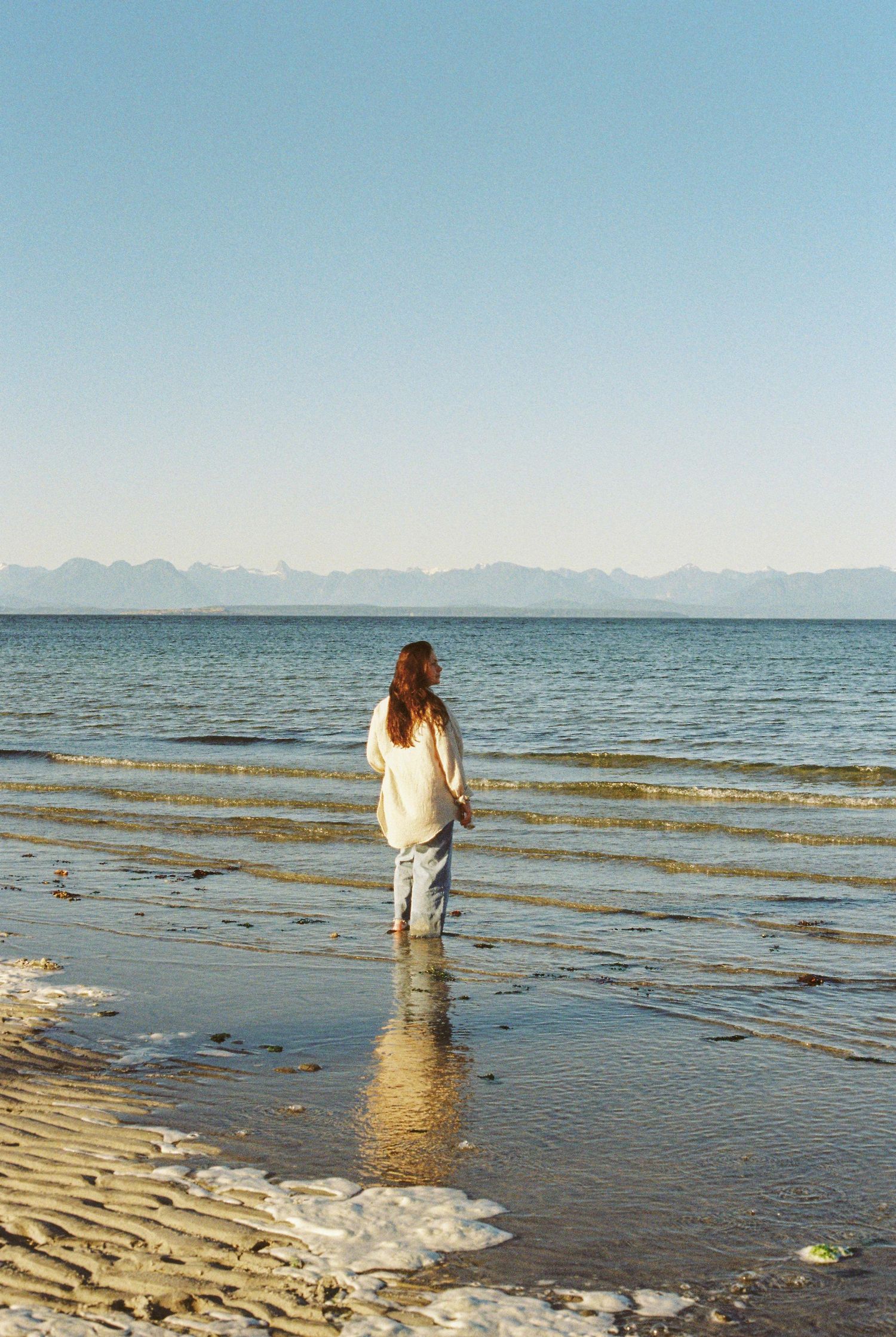 Woman in white top and jeans wading in shallow ocean water with mountains and clear blue sky in background.