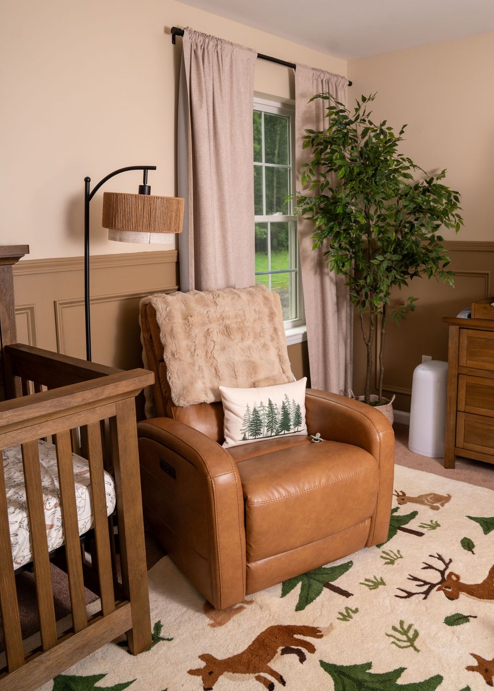 Nature-themed nursery with brown leather recliner, forest pillow, faux fur throw, woodland rug, wooden crib, and tall green plant.