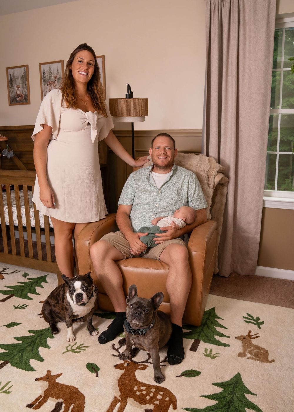 Family portrait in nursery with newborn baby, two French bulldogs on a deer-print rug, beside a white crib.
