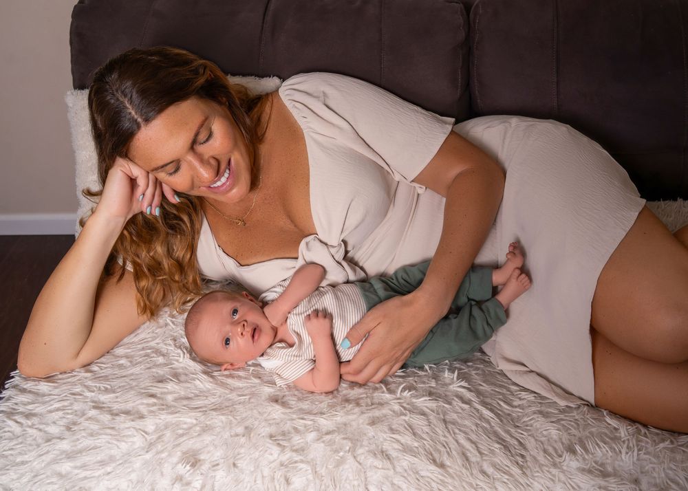 A smiling mother in a white dress lies on a fluffy rug, lovingly gazing at her newborn baby in green pants.