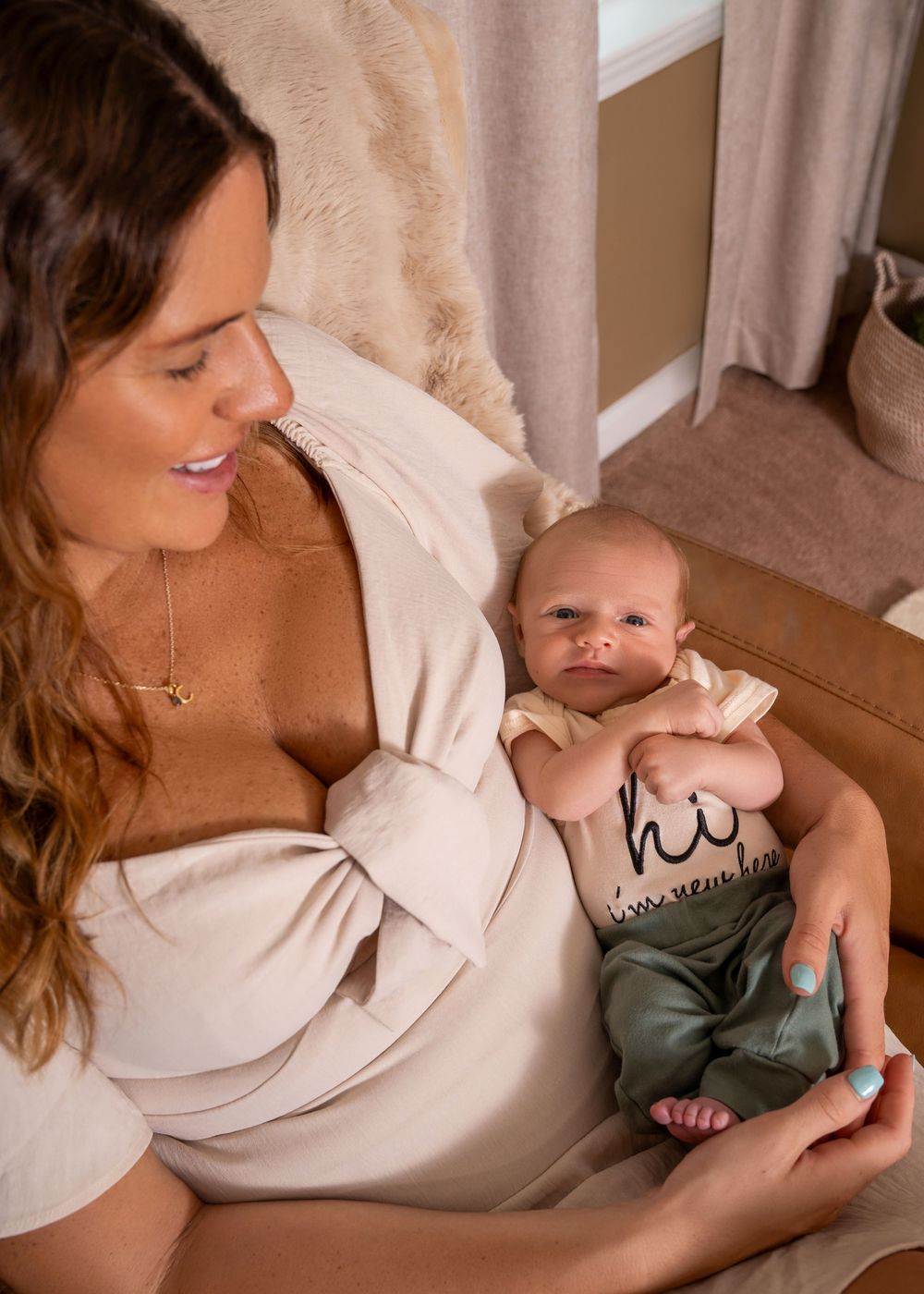 A young mother in a cream robe smiles down at her newborn baby wearing a green "hi" onesie.