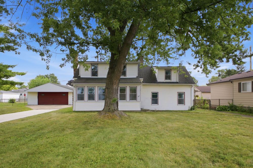 White two-story home with dormers, large shade tree in front yard, detached red garage, and well-maintained green lawn.