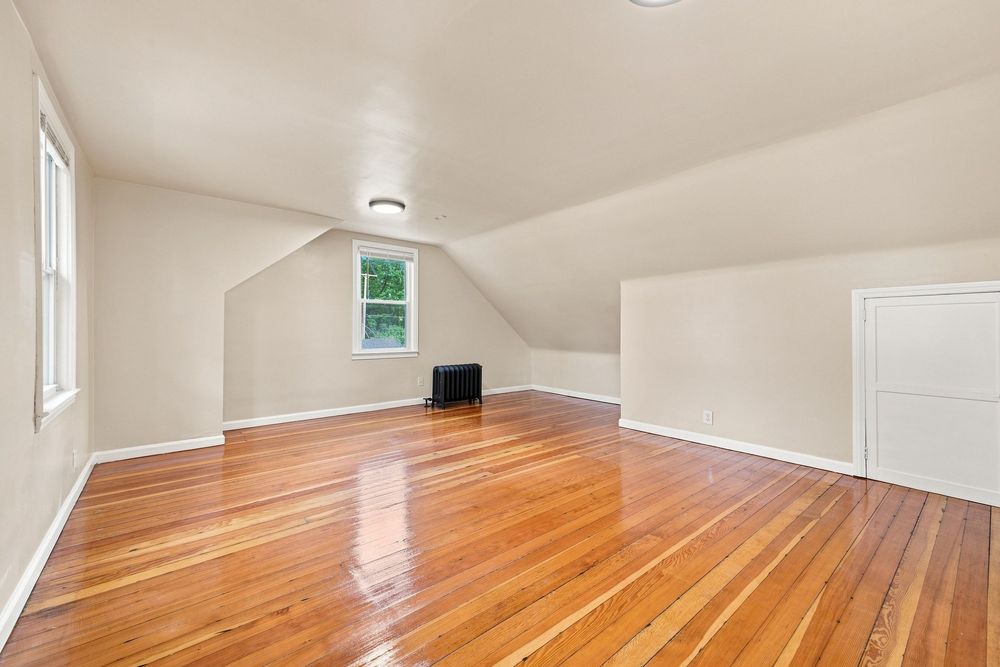Bright attic room with gleaming hardwood floors, white walls, sloped ceiling, and two windows letting in natural light.
