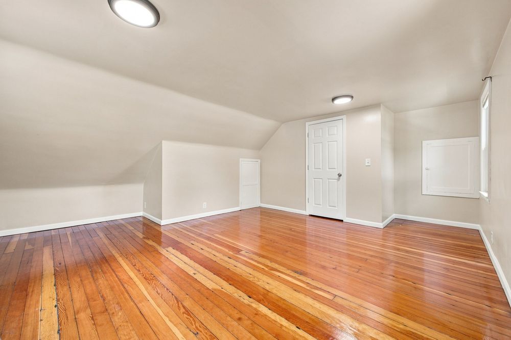 Spacious attic room featuring polished hardwood floors, vaulted ceiling, white walls, and a white door.