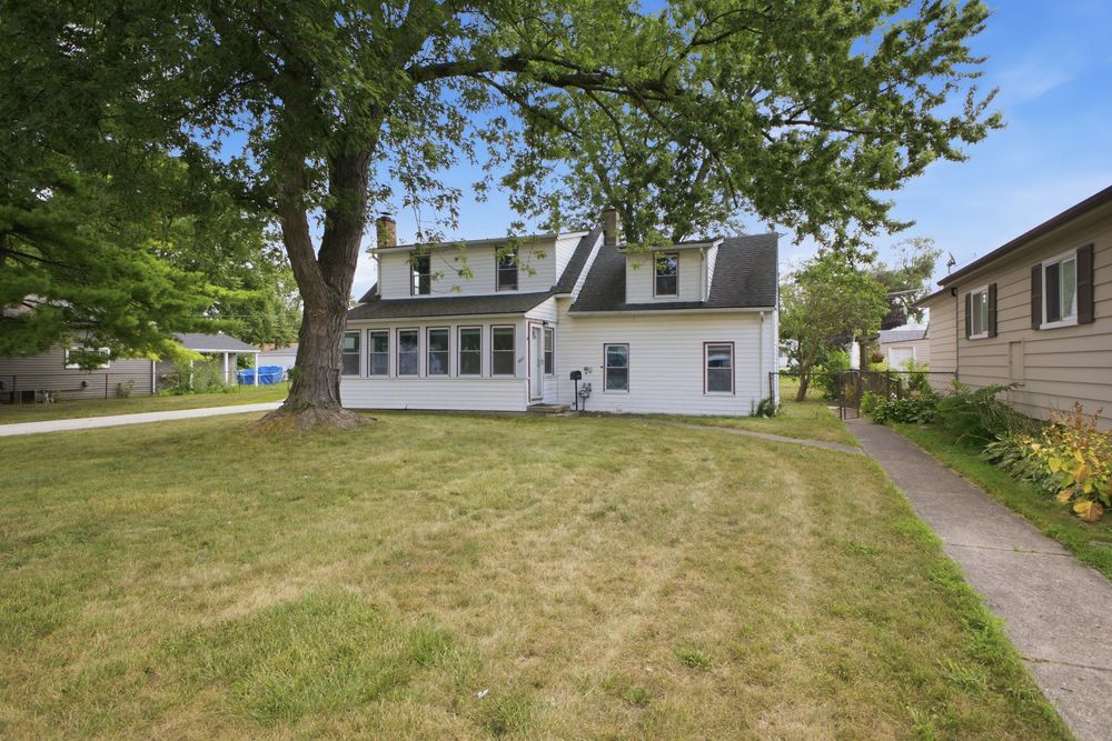 White two-story home with large shade tree, green lawn, and concrete driveway on a sunny day.
