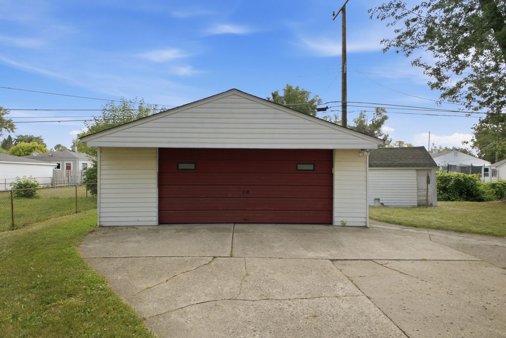 A detached two-car garage with a red door and white siding sits on a concrete driveway surrounded by green lawn.