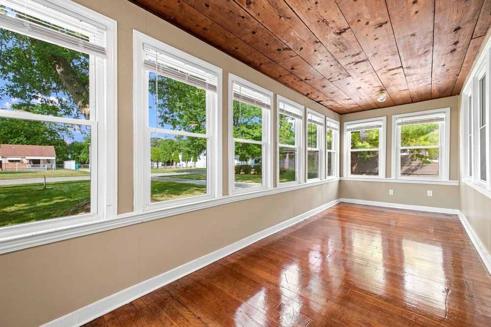 Bright sunroom with hardwood floors, wood plank ceiling, and large white-framed windows overlooking a lush green yard.