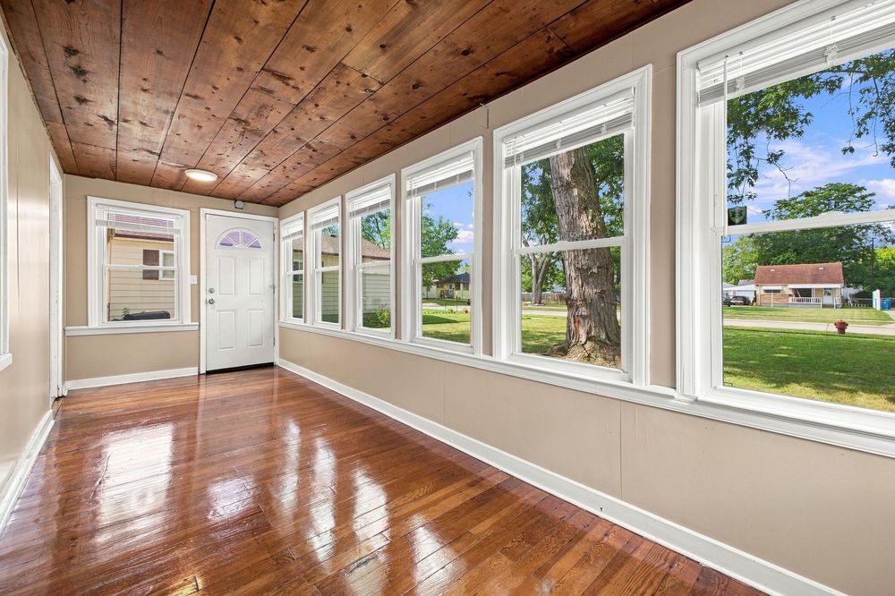 Bright sunroom with hardwood floors, wood ceiling, large windows overlooking lush green yard and mature trees.