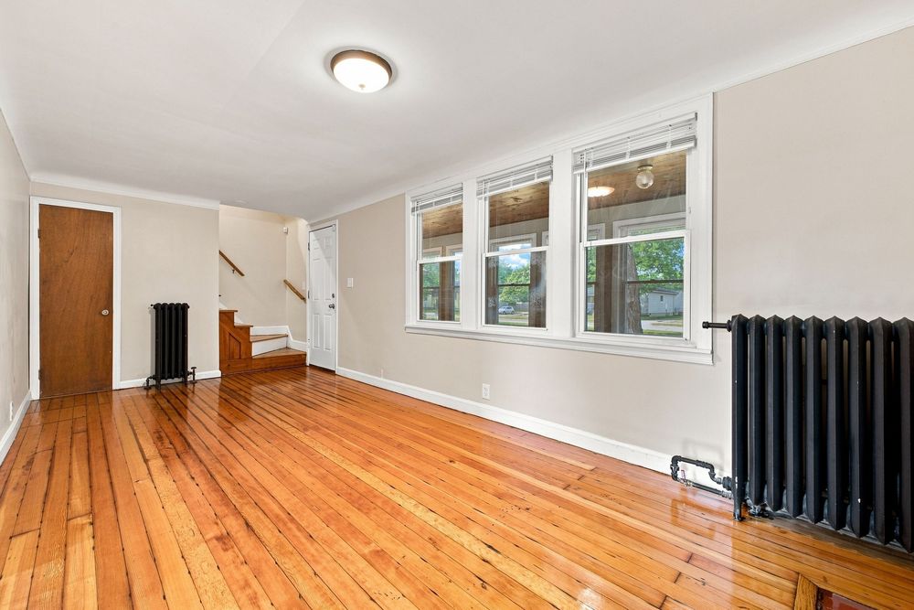 Bright living room with hardwood floors, white walls, large windows, black radiator, and staircase in a classic home.
