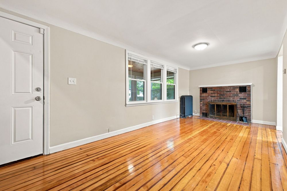 Bright living room with gleaming hardwood floors, crown molding, brick fireplace, and a white entry door with natural light.