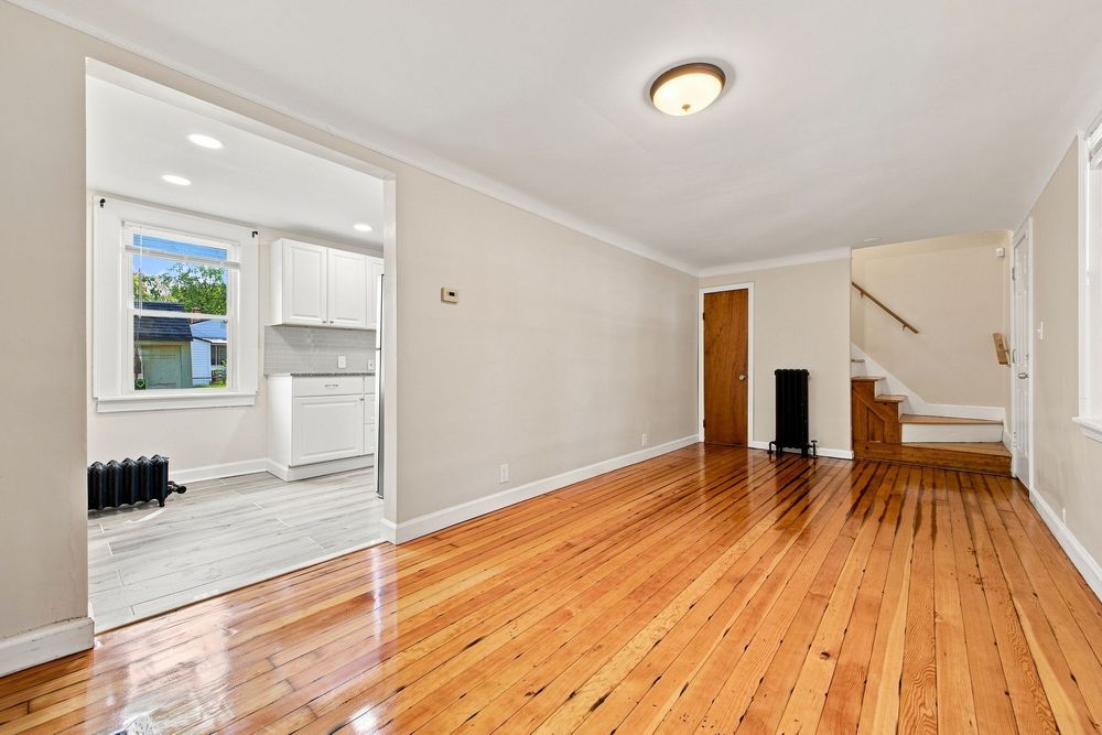 Bright living room with gleaming hardwood floors, crown molding, and open view to kitchen with white appliances.