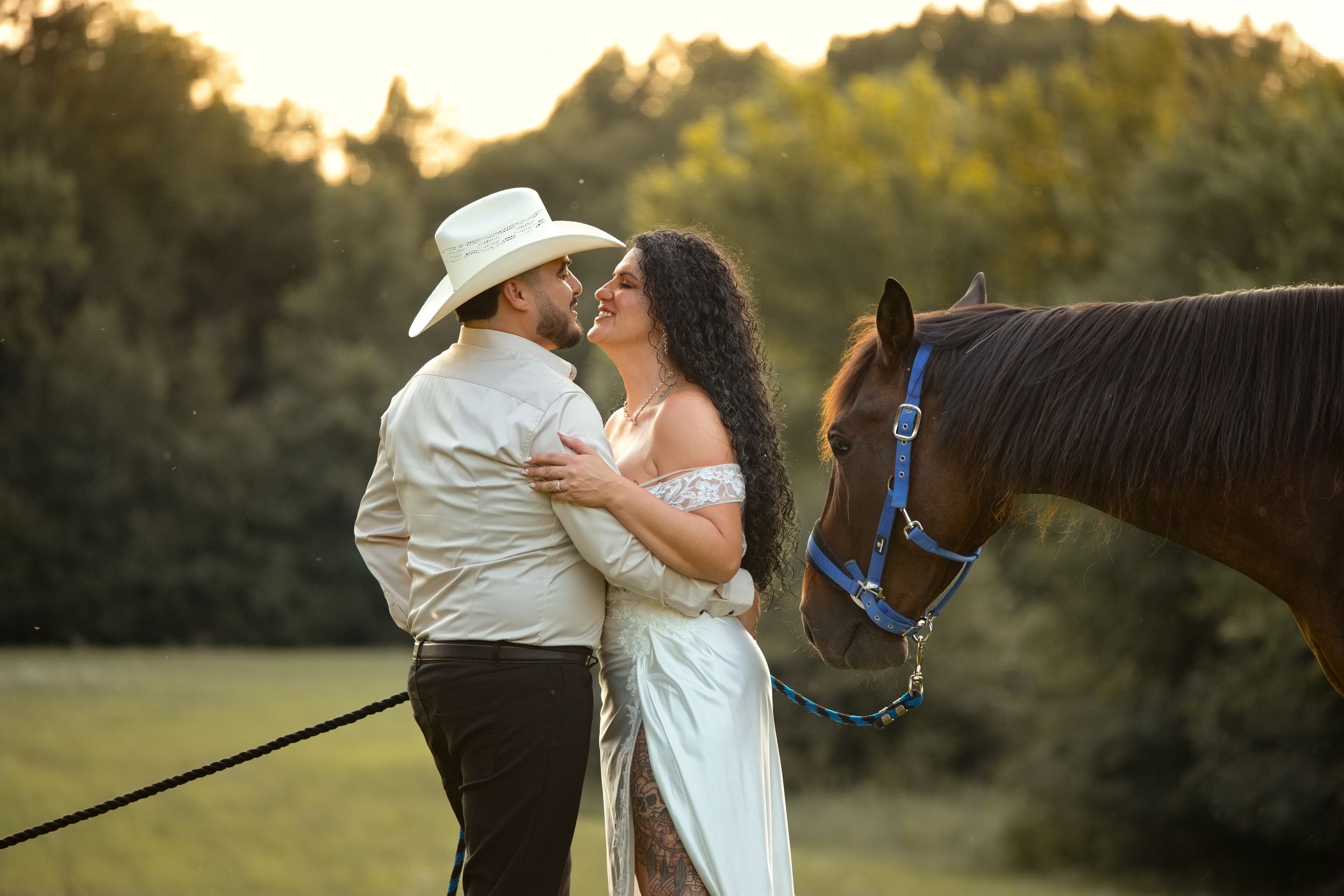 Couple sharing a romantic kiss in a golden field, man in cowboy hat and woman in white dress, with a brown horse nearby.