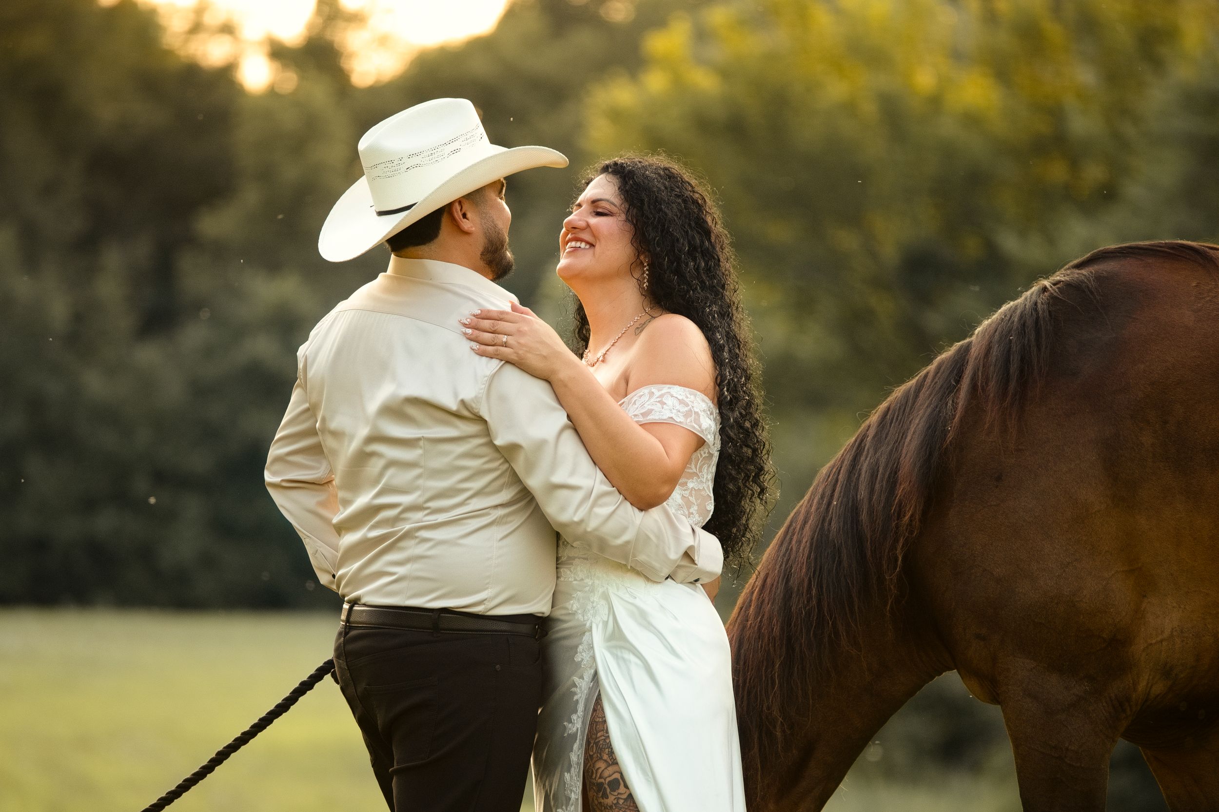 Couple in wedding attire embracing near a brown horse at golden hour in a lush green outdoor setting.