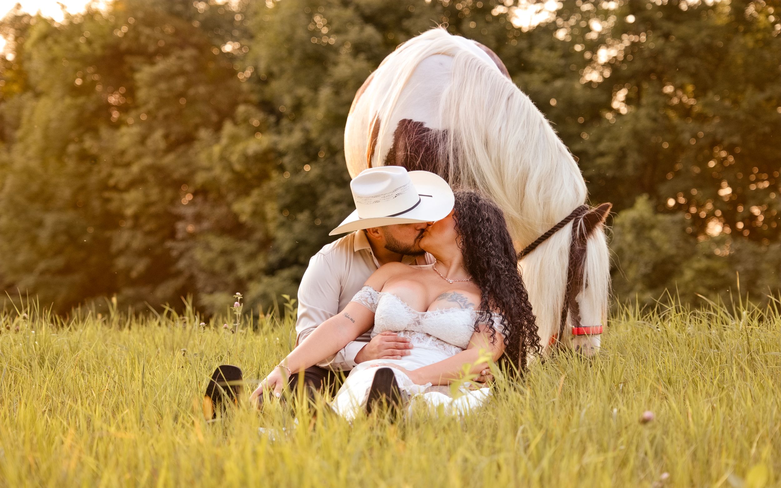 Couple sharing a kiss in a golden meadow with a white horse nearby, woman wearing a cowboy hat.