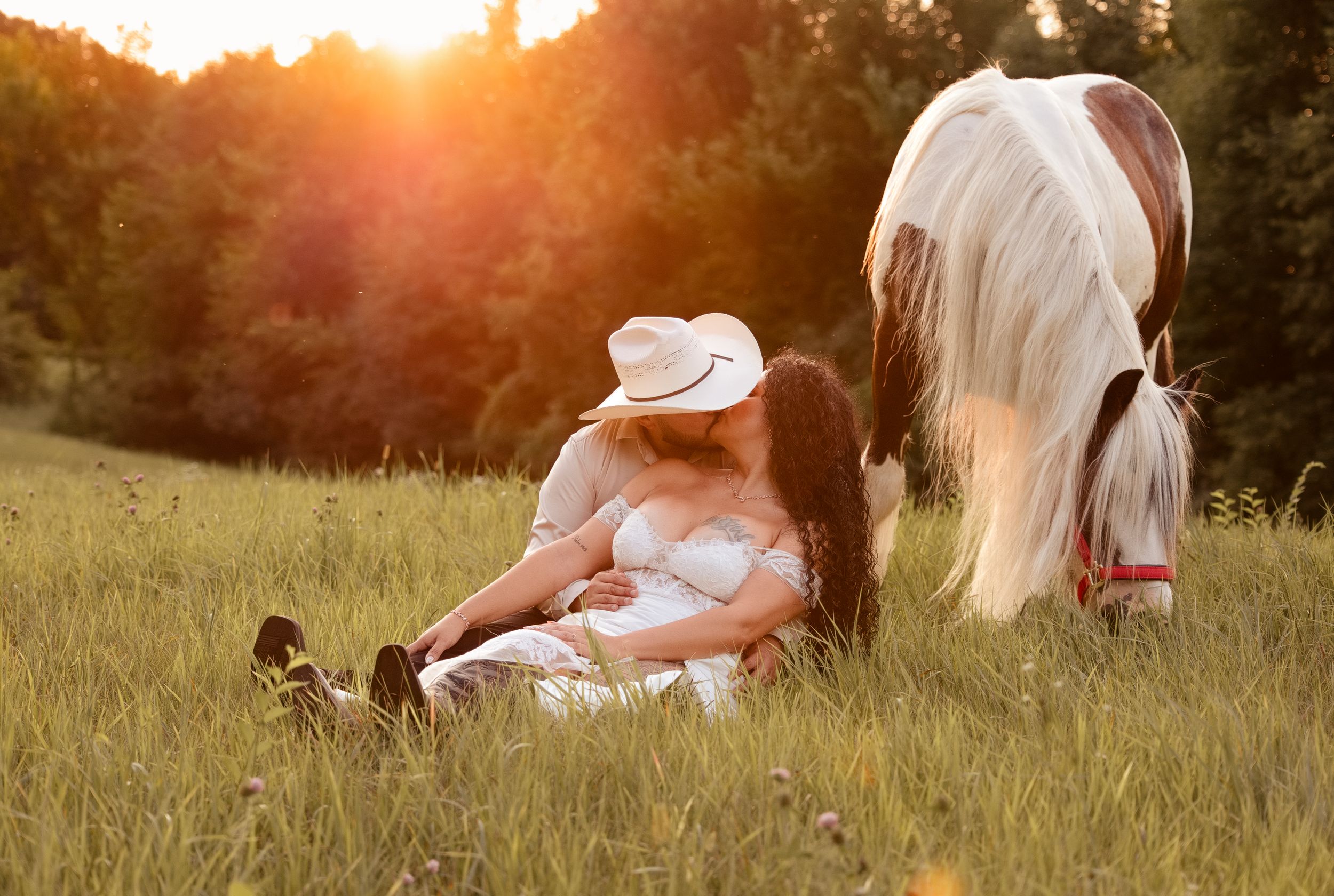 Woman in white dress and cowboy hat sits in golden sunset field beside a grazing white horse.