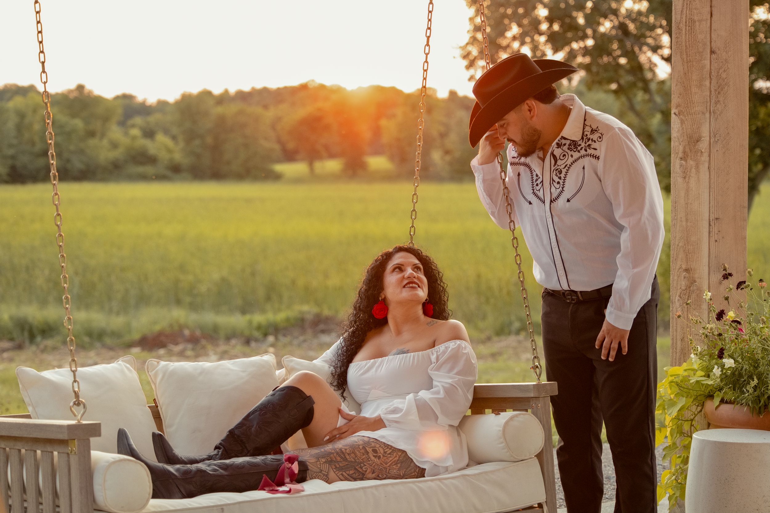 Couple in western attire on a porch swing at sunset, woman in white off-shoulder top, man in cowboy hat, green field backdrop.