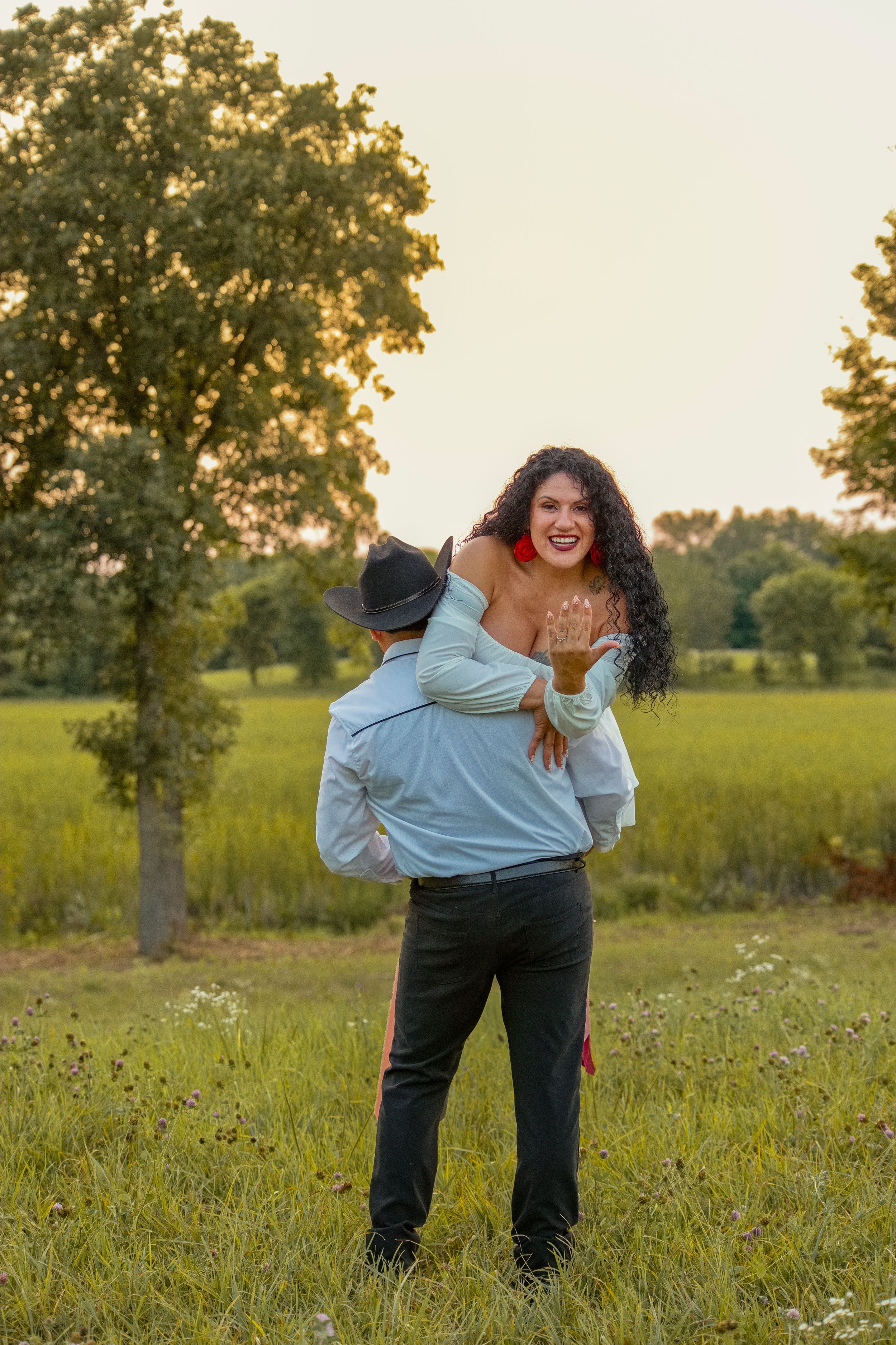 A man lifts a joyful curly-haired woman in a red dress outdoors in a green field at golden hour.