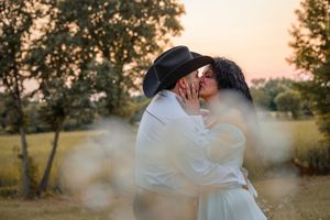 Couple sharing a romantic kiss at sunset in a field, man wearing a black cowboy hat and white shirt.