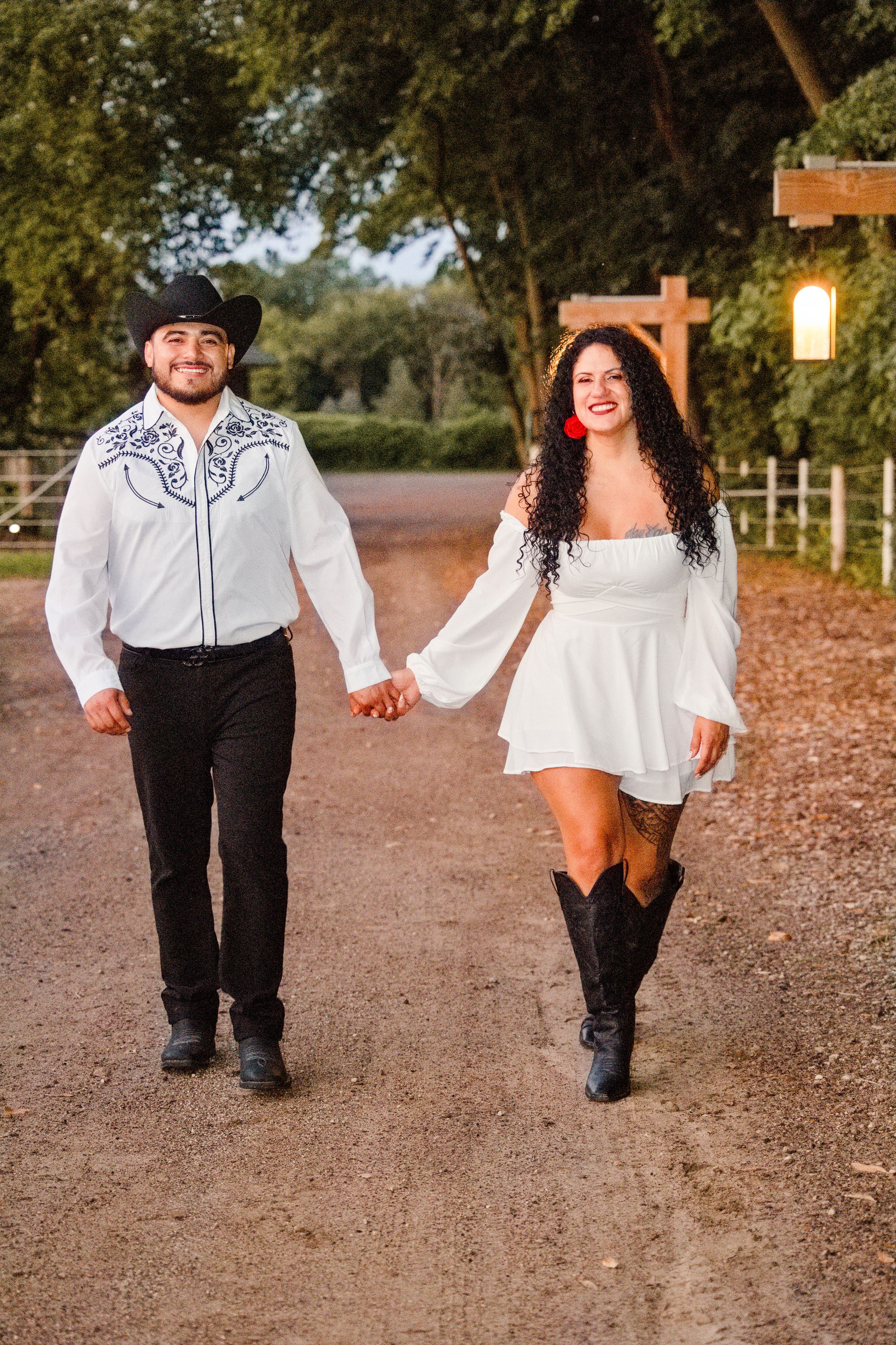 A couple in matching white outfits and cowboy boots walks hand-in-hand down a rustic path surrounded by lush green trees.