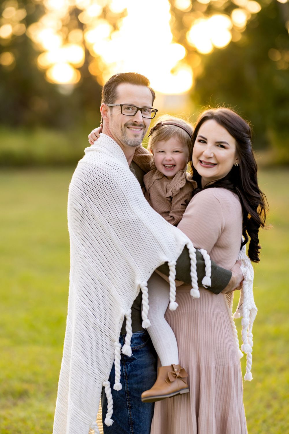 A happy family of three wrapped in a white blanket poses outdoors at golden hour in a sunlit green field.