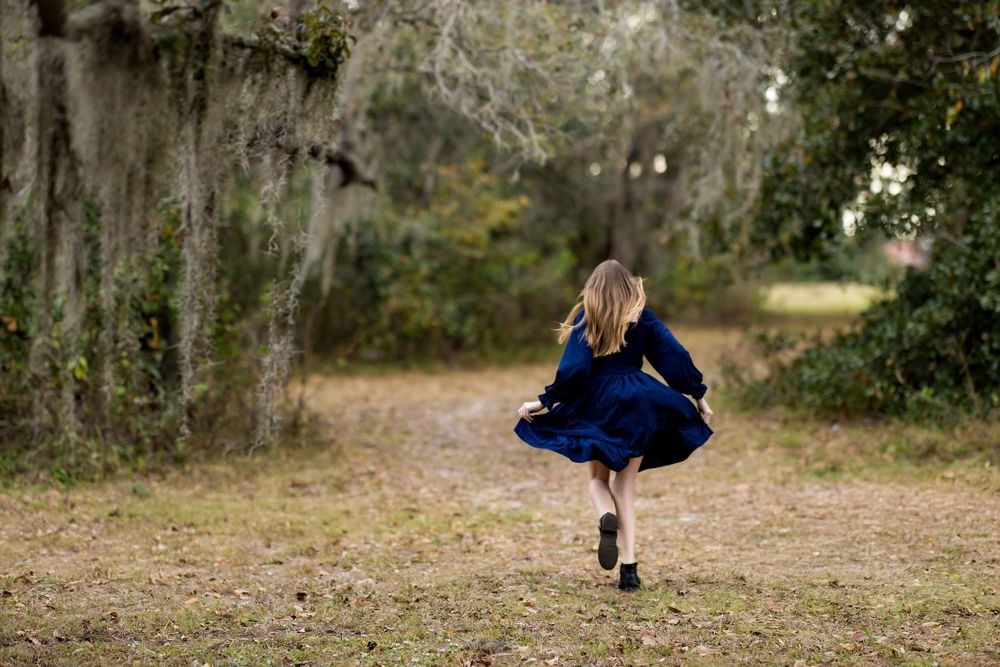 Girl in blue dress twirling on a dirt path surrounded by moss-draped trees in a lush outdoor setting.