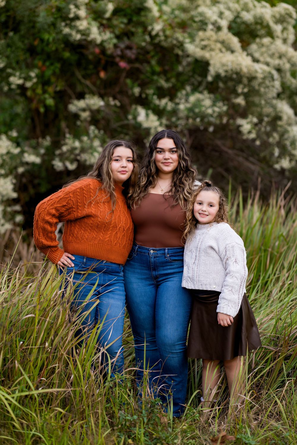 Three girls posing outdoors in tall grass with blooming bushes, wearing cozy sweaters and jeans in autumn colors.