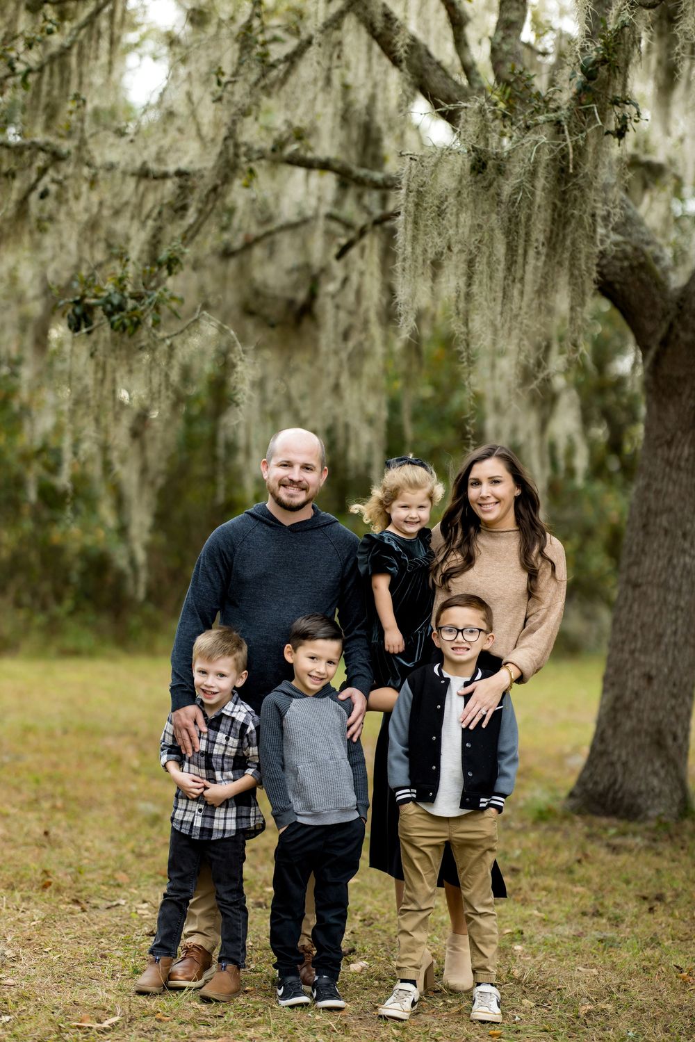 Family of six posing outdoors under Spanish moss-draped oak trees, with parents and four young children smiling together.