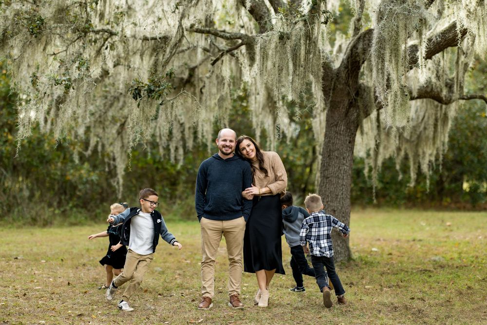 Family of four smiling under Spanish moss-draped oak tree in a lush green park, two young boys playing around their parents.