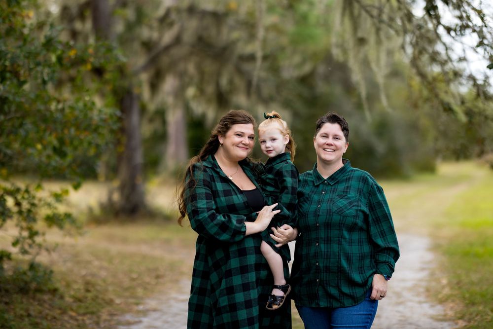 Two women in matching green plaid shirts pose outdoors on a tree-lined path, one holding a small child in a dark outfit.