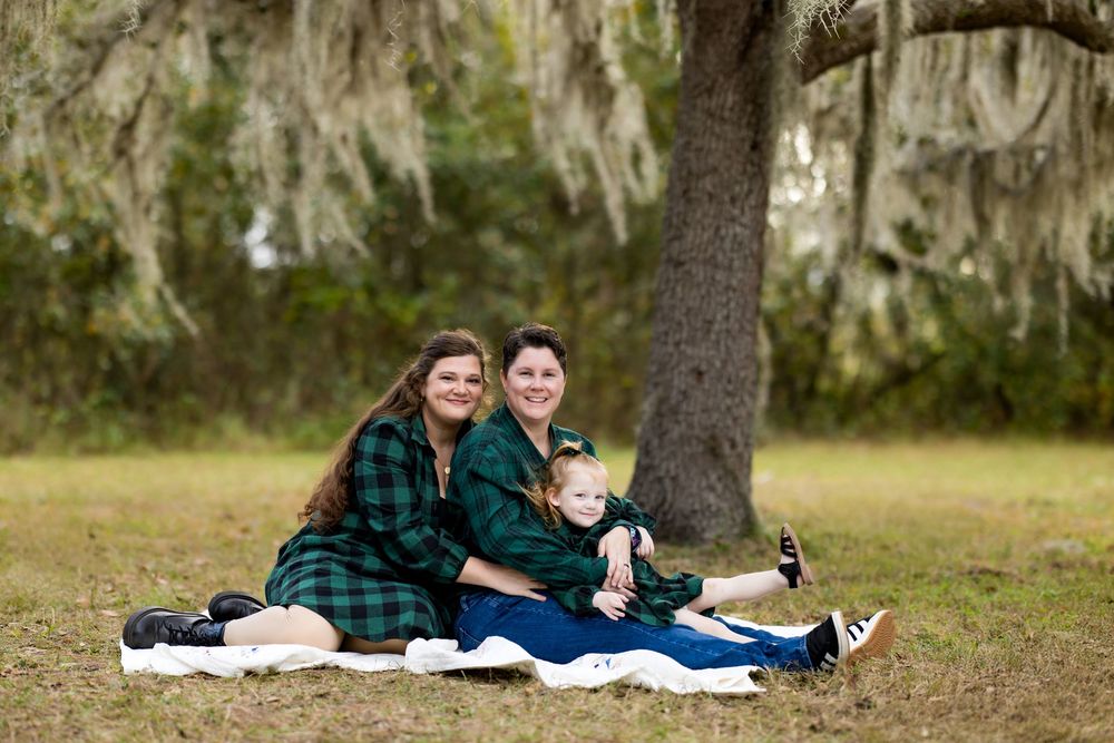 Family of three in matching green plaid outfits sitting on blanket under moss-covered tree outdoors.
