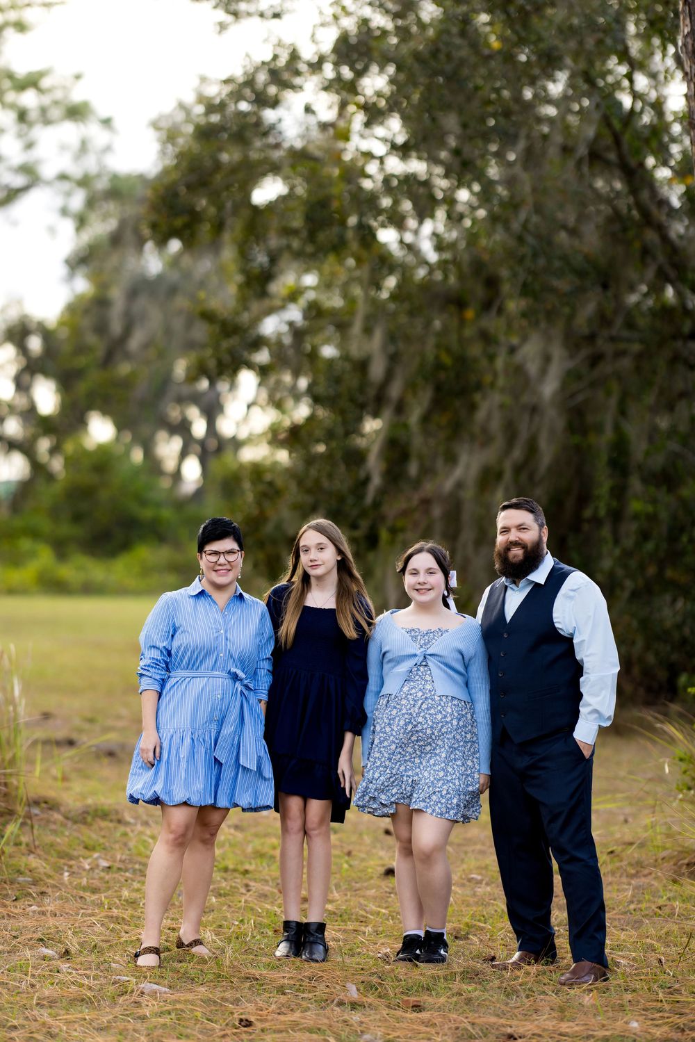 Family of four posing outdoors in coordinating blue outfits on a grassy path with lush green trees behind them.
