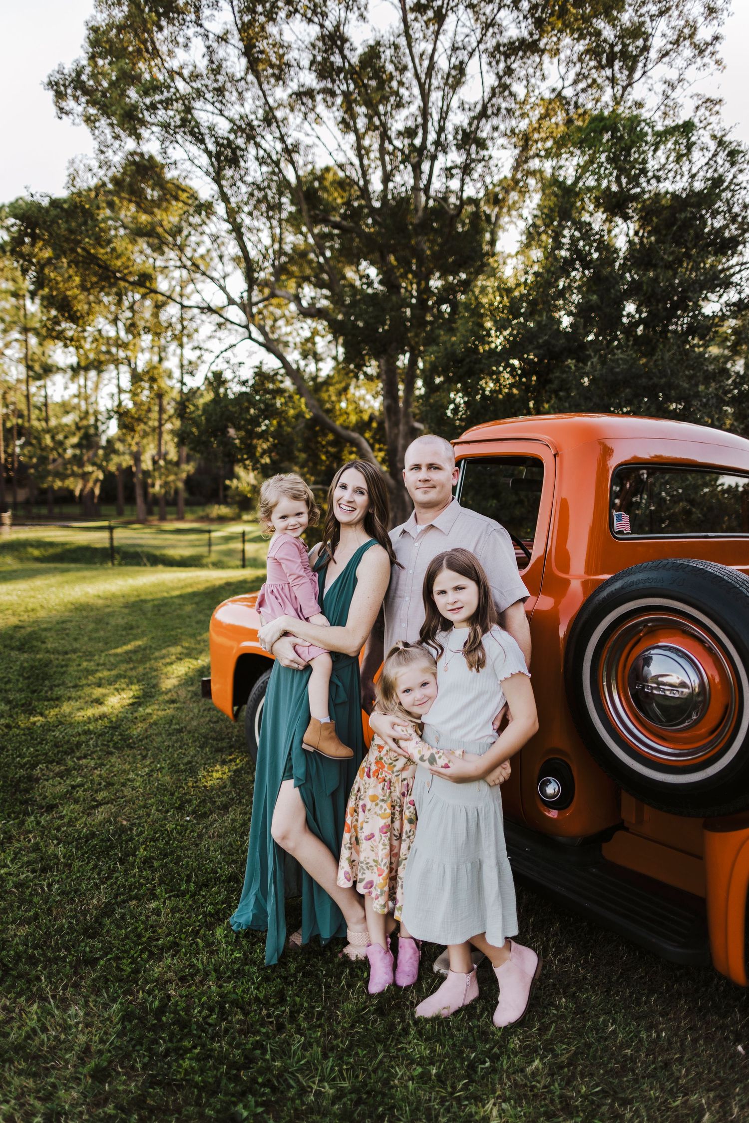 Family of five posing beside a classic orange vintage truck on a sunny day in a green park setting.