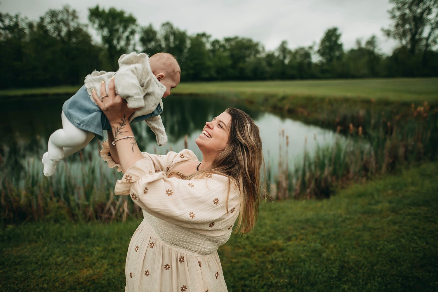 A joyful mother in a floral dress lifts her smiling baby outdoors near a serene pond with lush greenery.