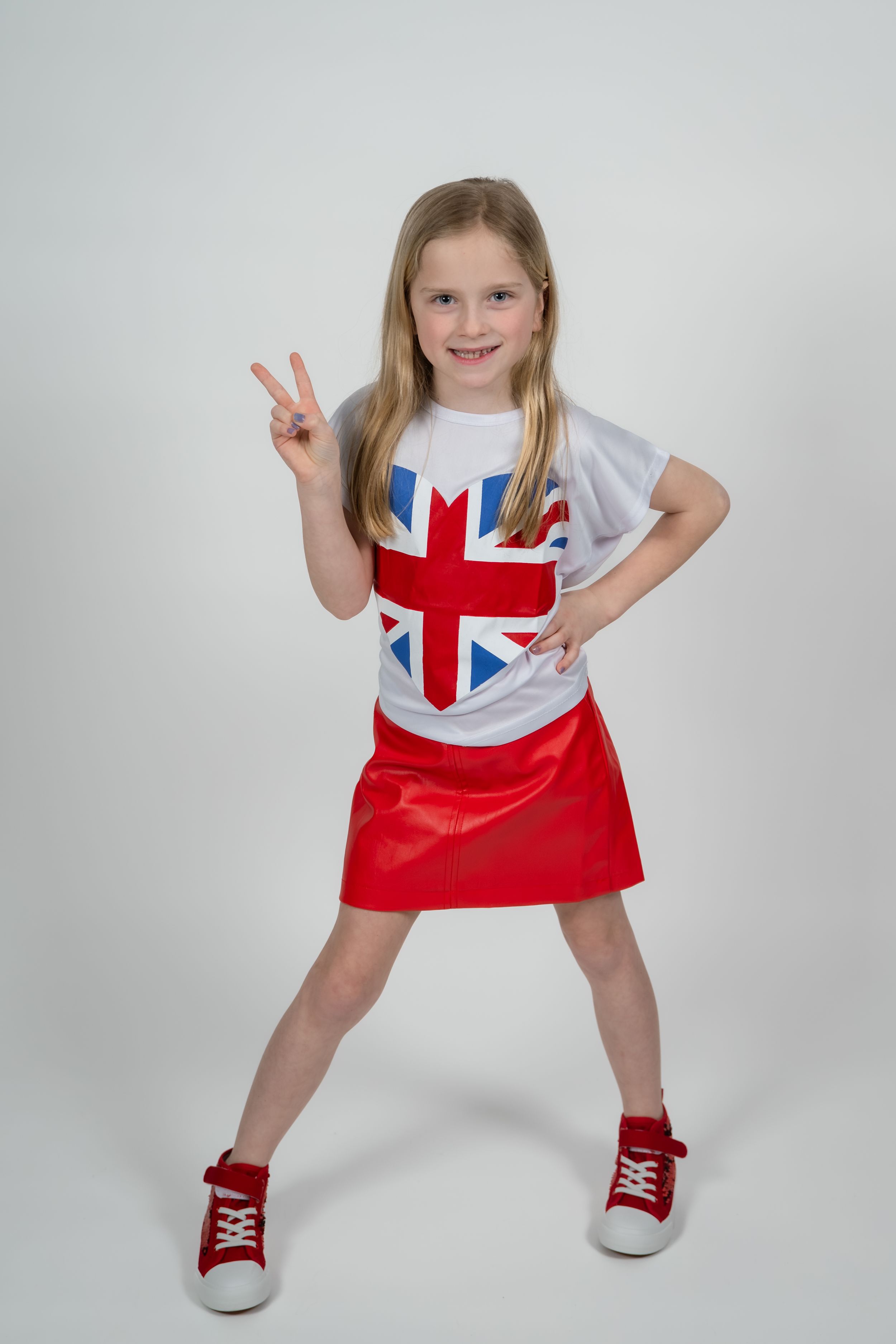 Young girl in Union Jack t-shirt and red skirt making peace sign, smiling confidently in studio.