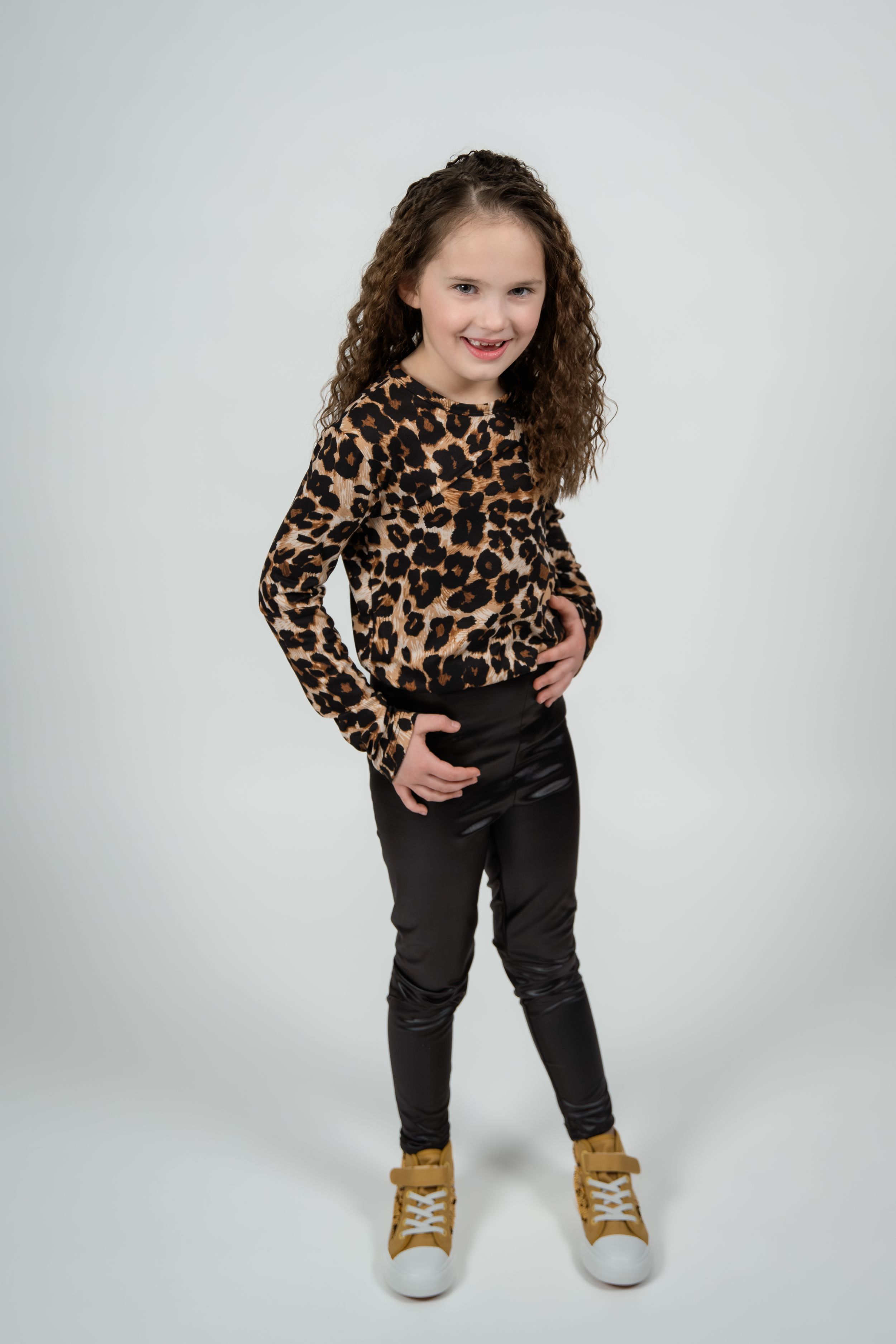Young girl in leopard print top, black shiny leggings, and gold sneakers posing confidently in a studio.
