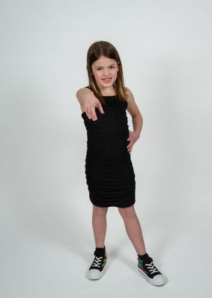 Girl in black ruched dress and colorful sneakers pointing at camera on white background.