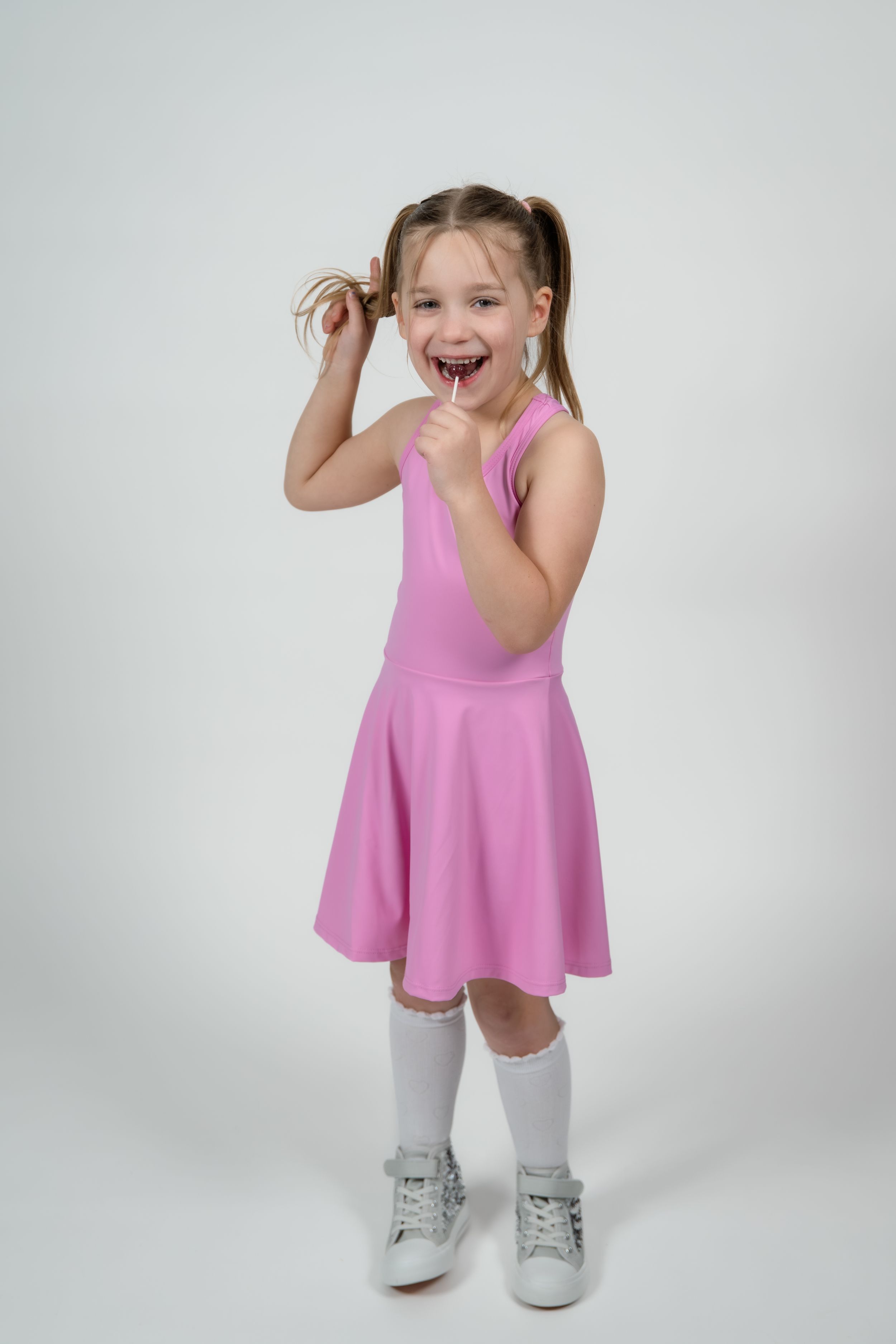 Happy little girl in pink dress and white knee socks holding a lollipop, smiling with pigtails on white background.