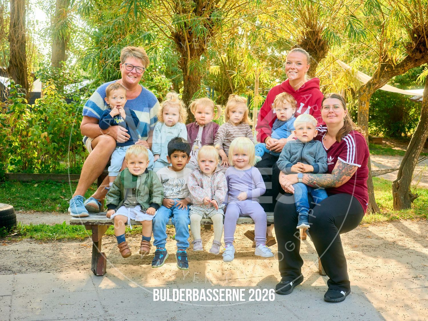 Family group photo with adults and children posing on a bench outdoors, labeled Bulderbasserne 2026.