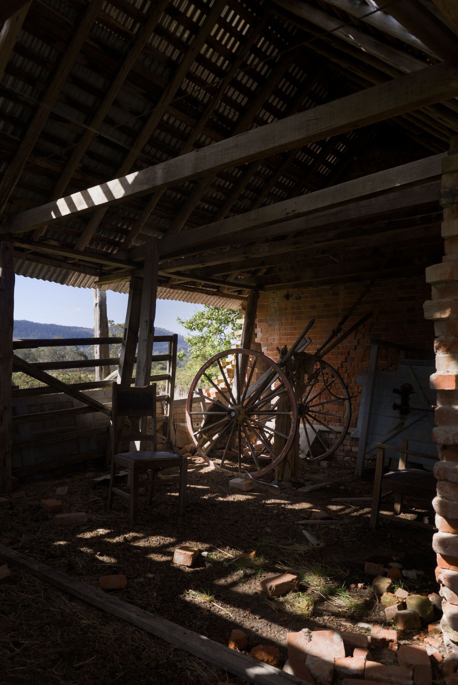Rustic abandoned barn interior with large wooden wagon wheels leaning against brick wall, scattered bricks on dirt floor.