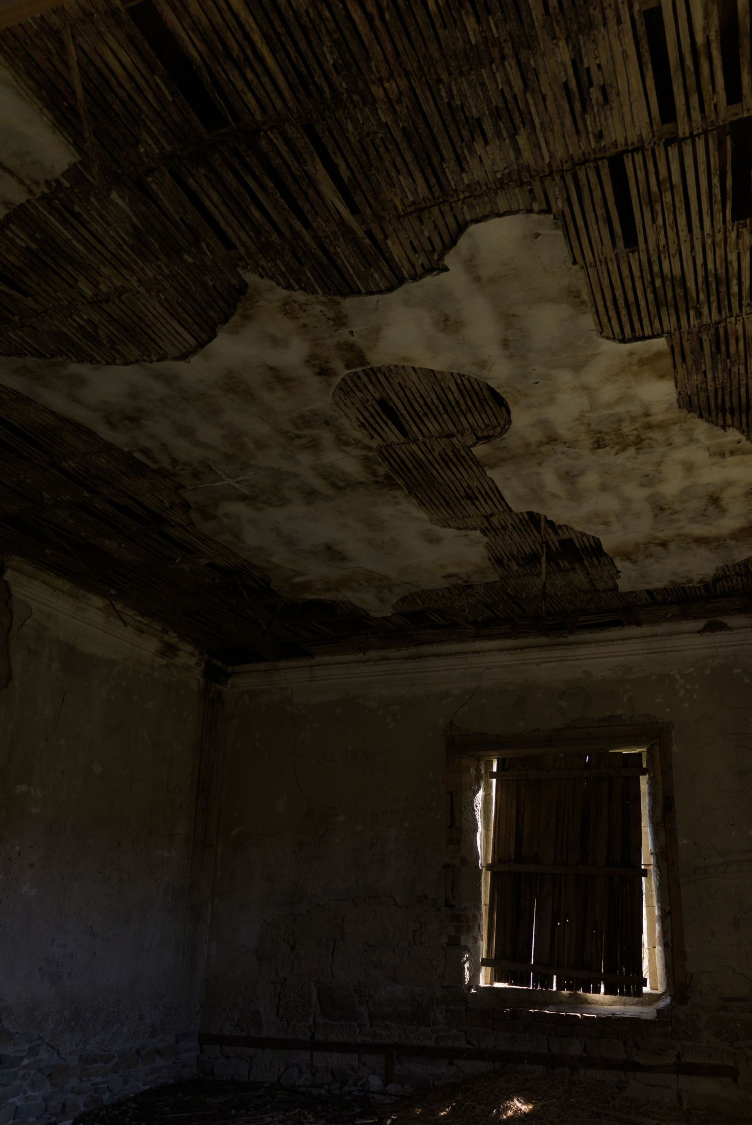 Abandoned room with deteriorating ceiling, exposed wooden laths, peeling plaster, and a broken window in a dark, decayed building.