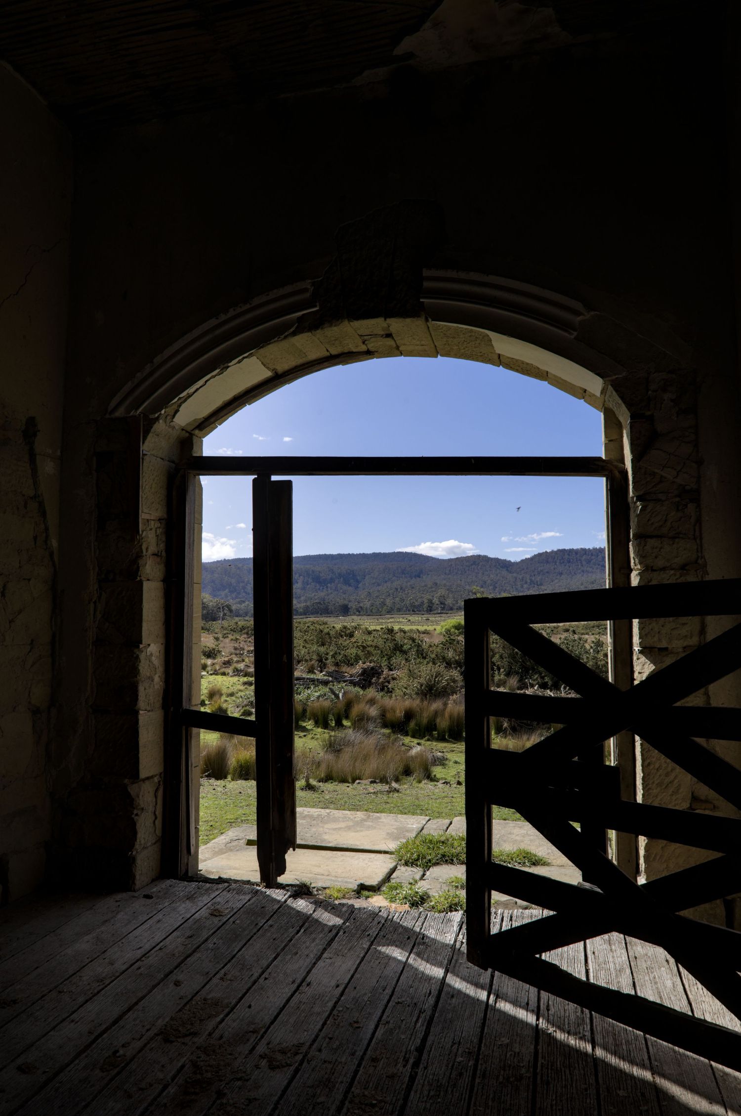 View through a stone archway with wooden gate revealing a scenic valley landscape under blue skies.