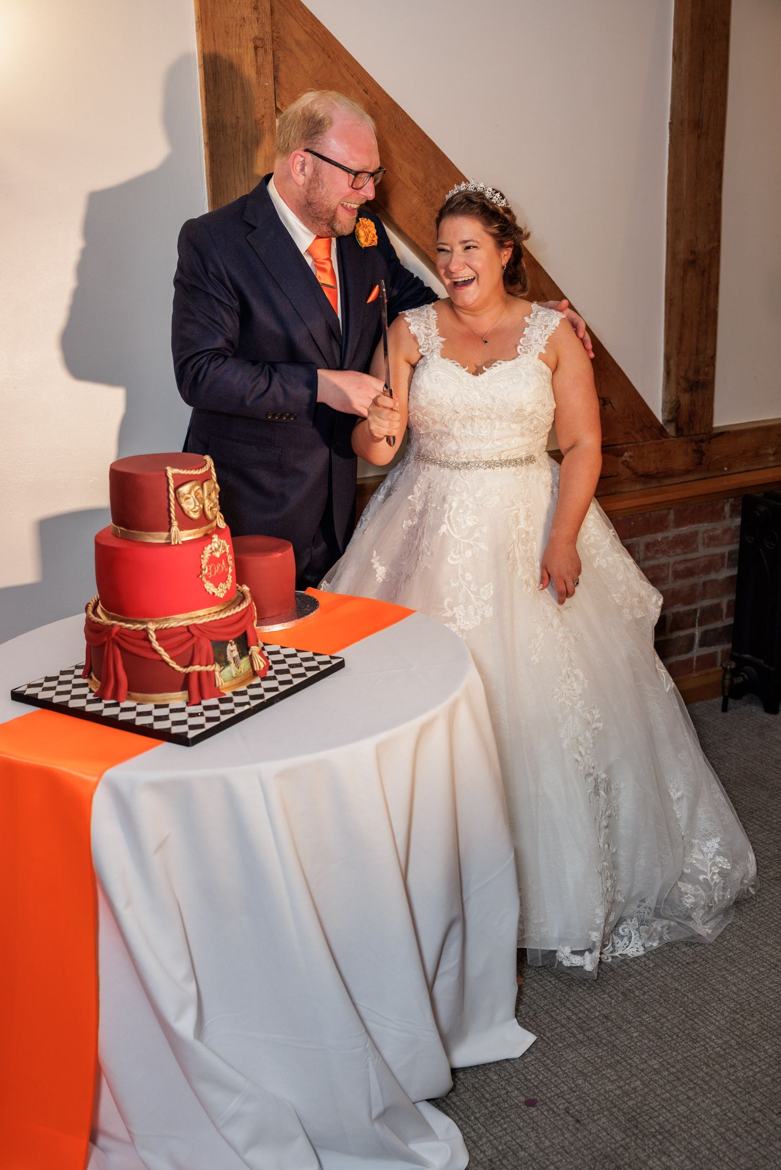 Bride and groom laughing together while cutting a red tiered wedding cake at their wedding reception.
