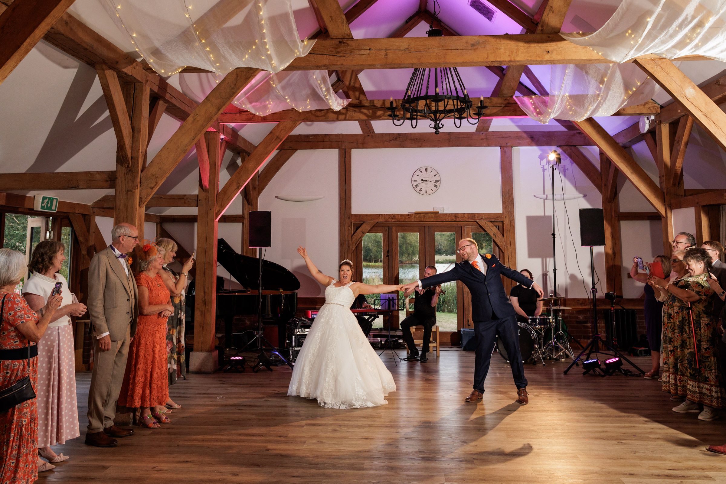 Bride and groom share first dance in rustic barn wedding venue with wooden beams and guests watching.