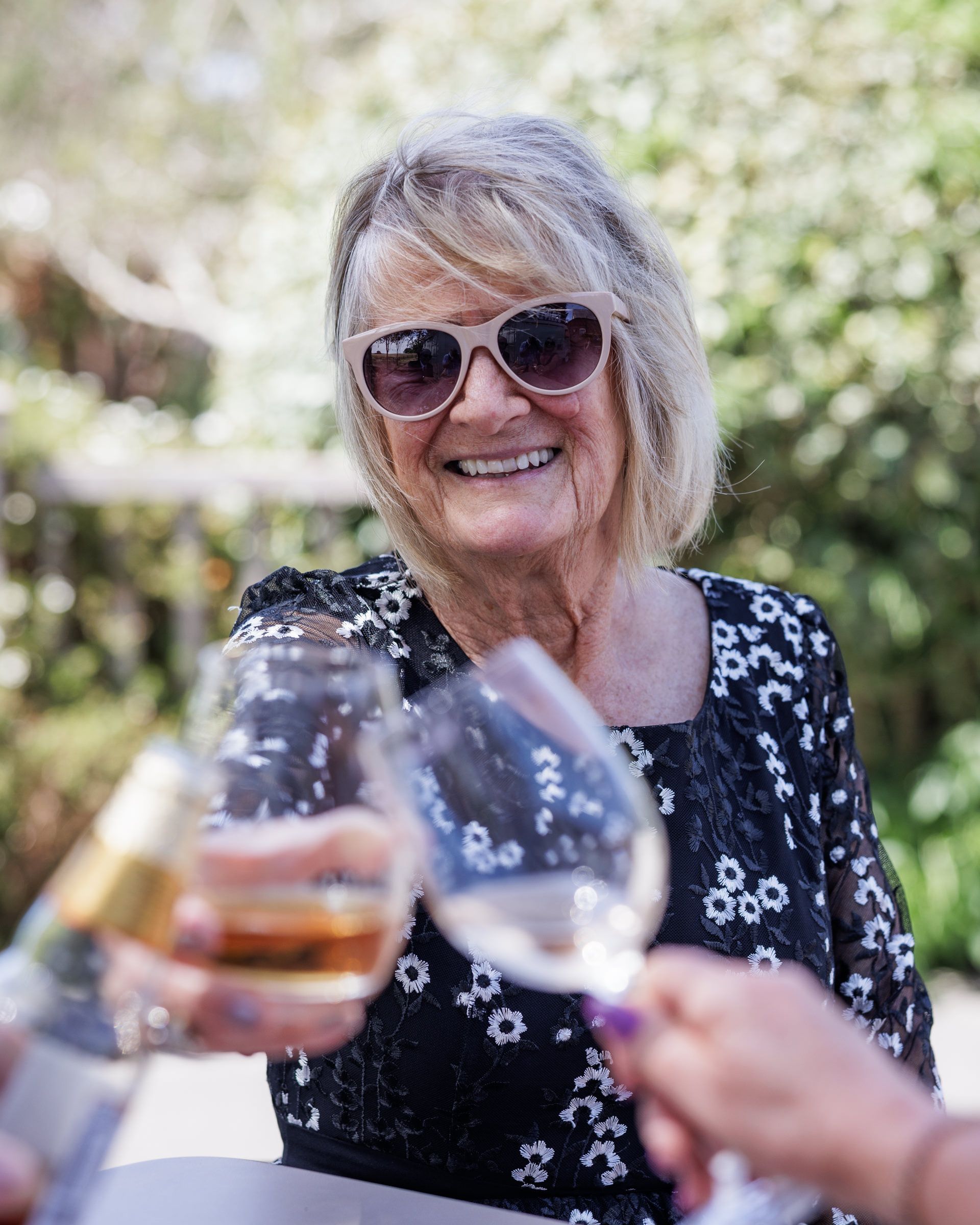 Smiling older woman in sunglasses toasting wine glasses outdoors at a sunny garden gathering.