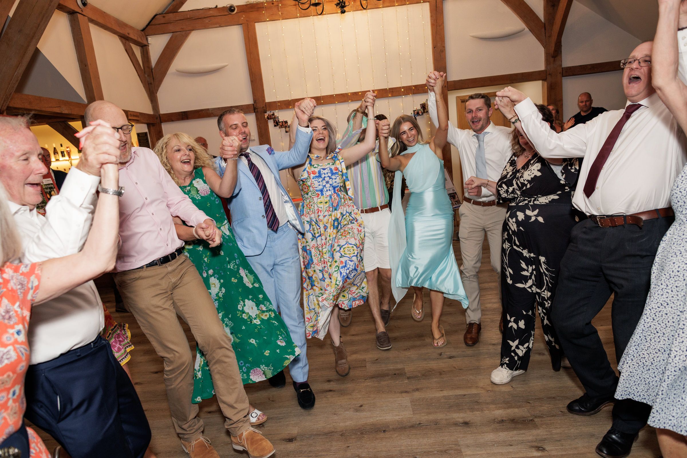 Guests dancing joyfully at a wedding reception in a rustic venue with wooden beams and warm lighting.