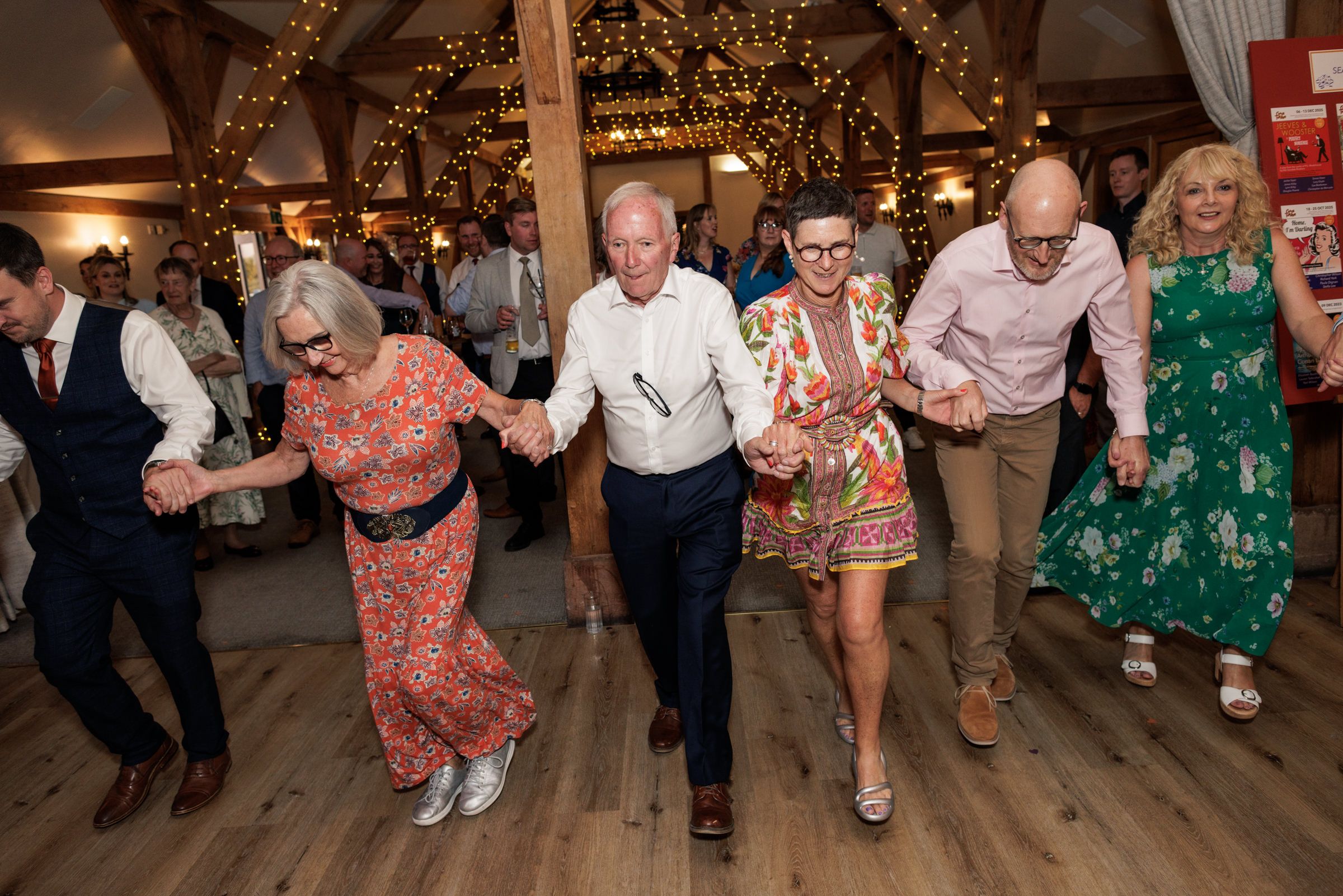 Guests dancing in a line at a wedding reception in a warmly lit barn venue decorated with fairy lights.